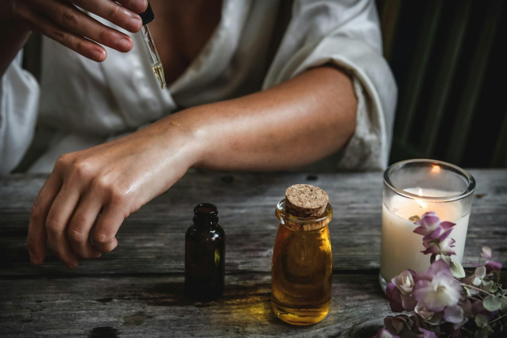 person pouring oil on their forearm with a dropper with oil bottles, a candle and flowers in the foreground.