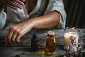 person pouring oil on their forearm with a dropper with oil bottles, a candle and flowers in the foreground.